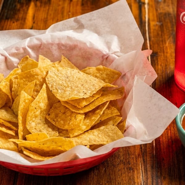 A red basket of tortilla chips with a side of salsa and a Coca-Cola on a wooden table.