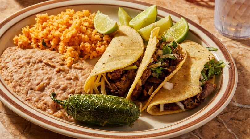 Three tacos with rice, refried beans, a grilled jalapeño, and lime wedges on a plate.