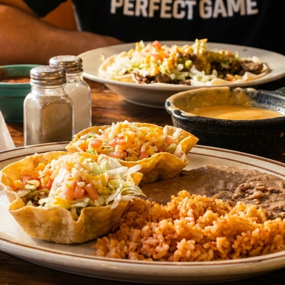 Two taco salads in edible bowls with rice and beans, a bowl of queso, and chips on a wooden table.