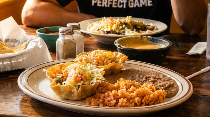 Two taco salads in edible bowls with rice and beans, a bowl of queso, and chips on a wooden table.